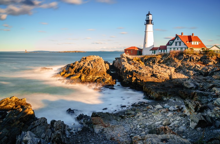 A red and white lighthouse sits on the water of a New England coastline.